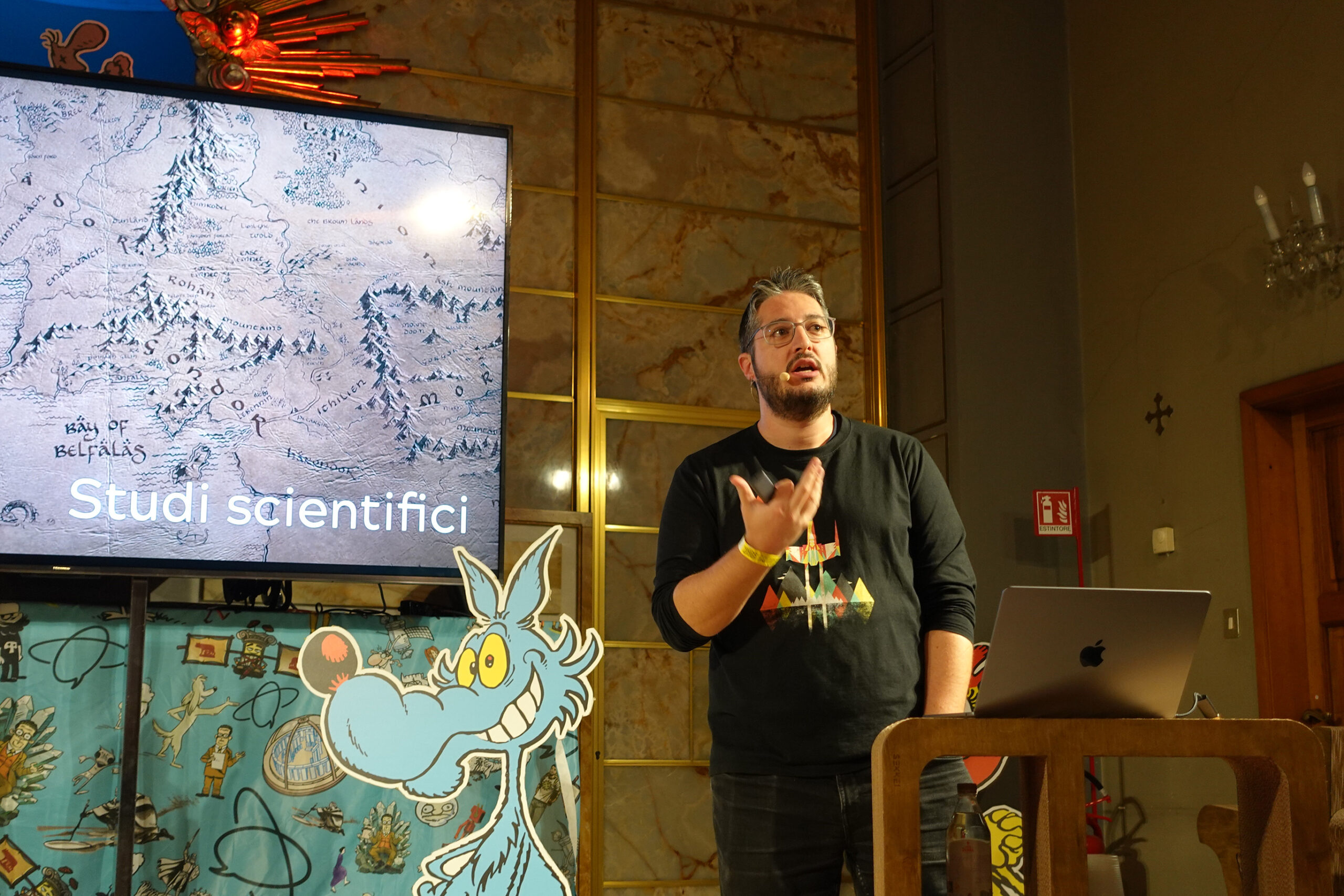 Al centro c'è un uomo con occhiali e capelli corti brizzolati. Parla muovendo la mano destra, la sinistra è in tasca. Di fronte a lui c'è un computer Apple appoggiato a un tavolo di cartone: sta proiettando una presentazione che si intravede nello schermo al plasma alle spalle dell'uomo. Nello schermo si vede una porzione della mappa della Terra di Mezzo del Signore degli Anelli di Tolkien e la scritta "Studi scientifici". L'uomo e lo schermo sono all'interno di una stanza con luci soffuse: completano l'arredo una sagoma di LUPO ALBERTO, una parete in finto marmo e un candelabro alla parete.